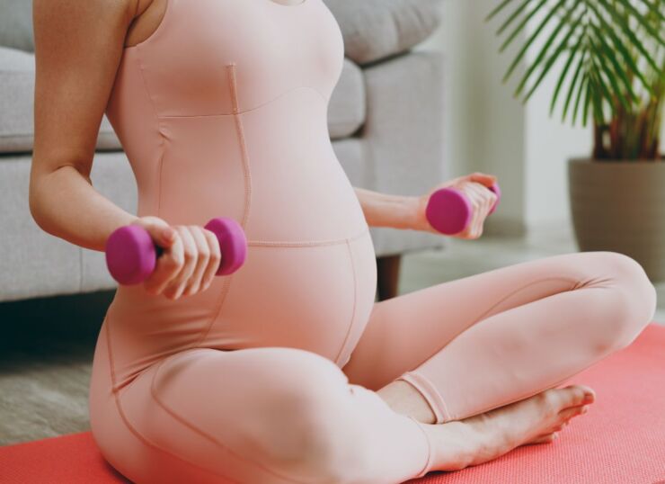 prikaz trudnice koja sedi na podlozi za jogu i podiže male tegove za ruke